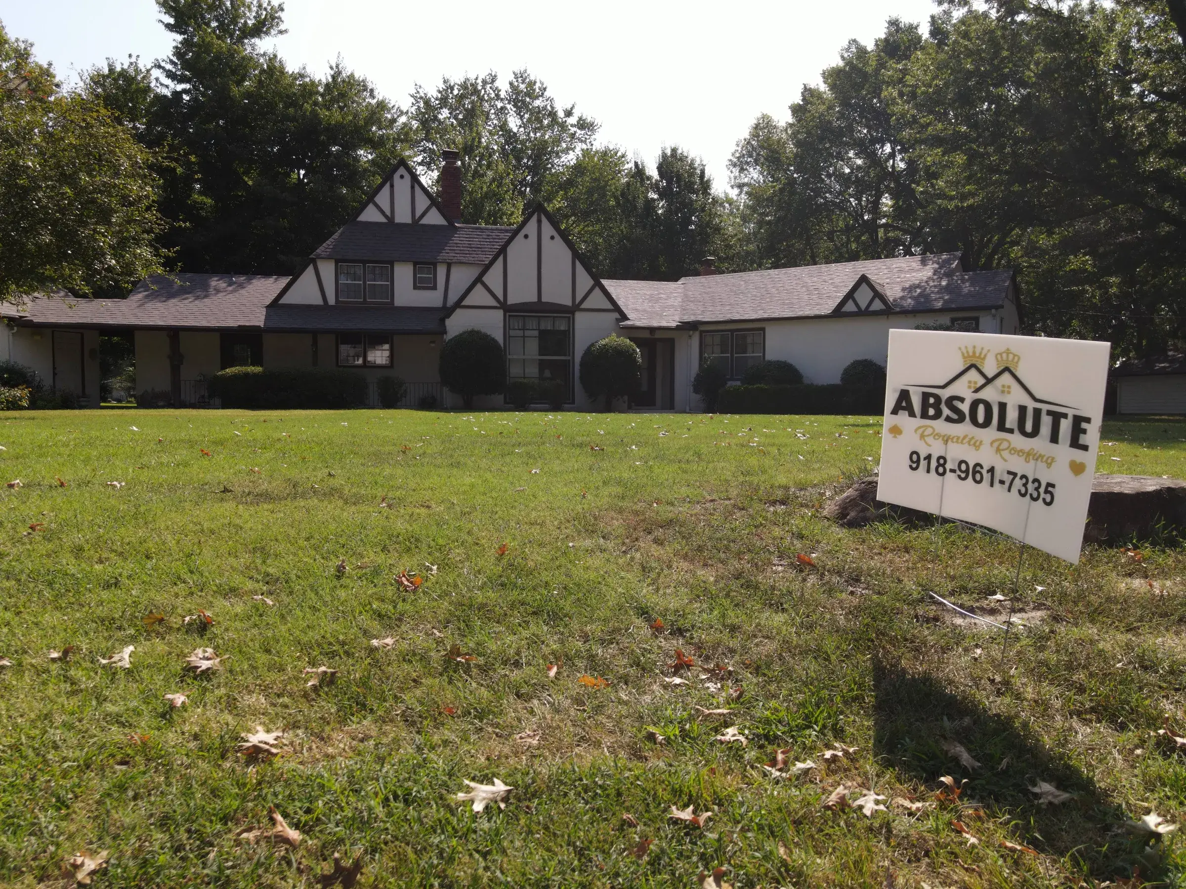 An Absolute Royalty Roofing yard sign on the lawn of a large Tudor-style home with a gray shingle roof.
