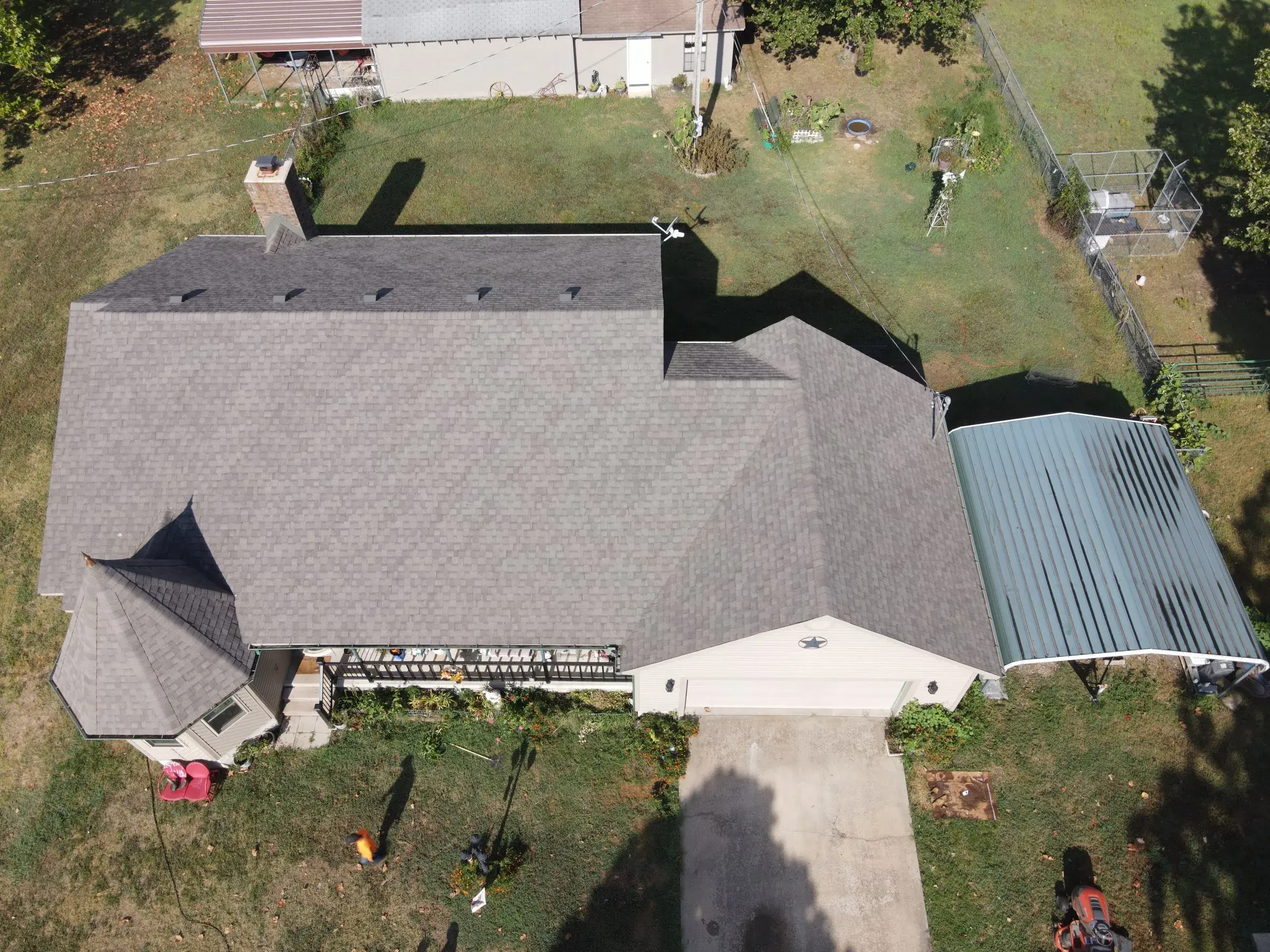 Overhead drone view of a house with a light gray shingle roof and ridge vents, next to a green metal carport roof.