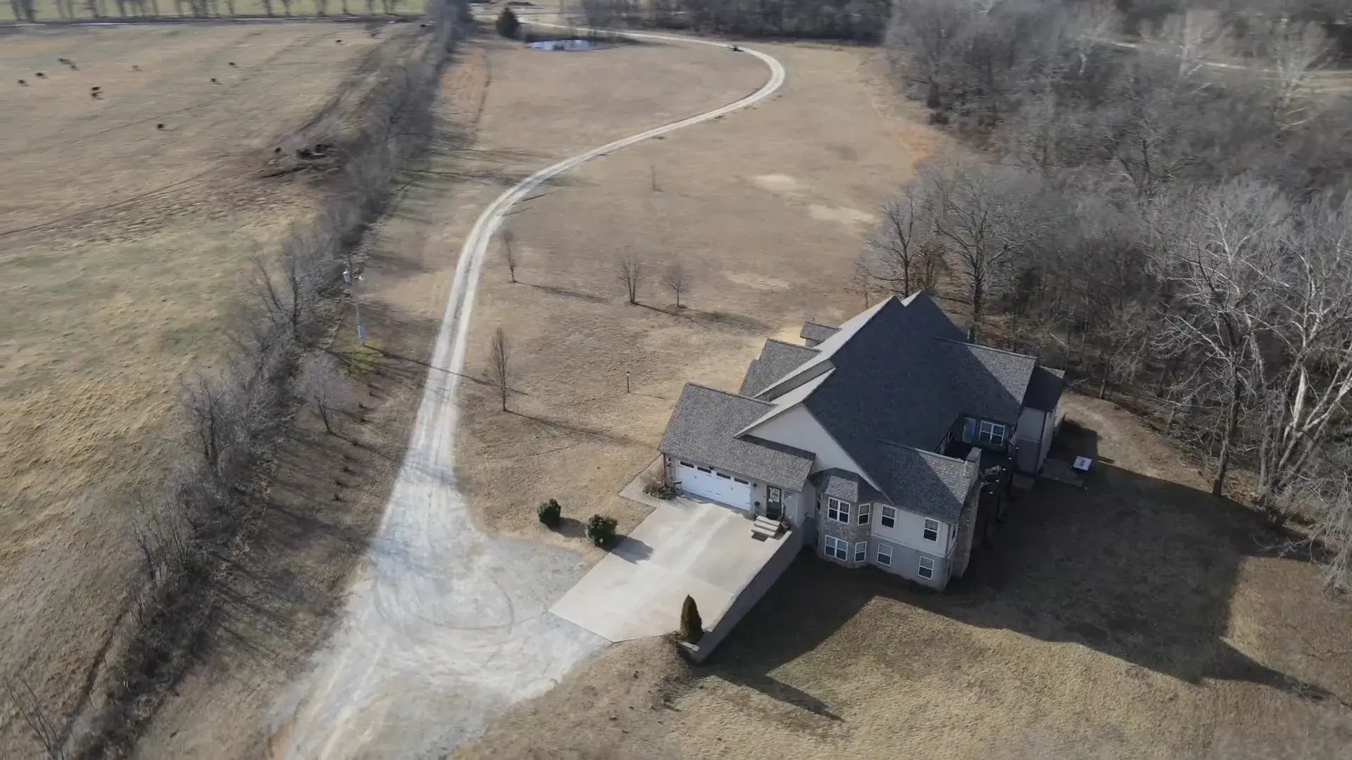 Wide aerial view of a large two-story home with a gray shingle roof at the end of a long gravel driveway, surrounded by dormant winter pasture and bare trees.
