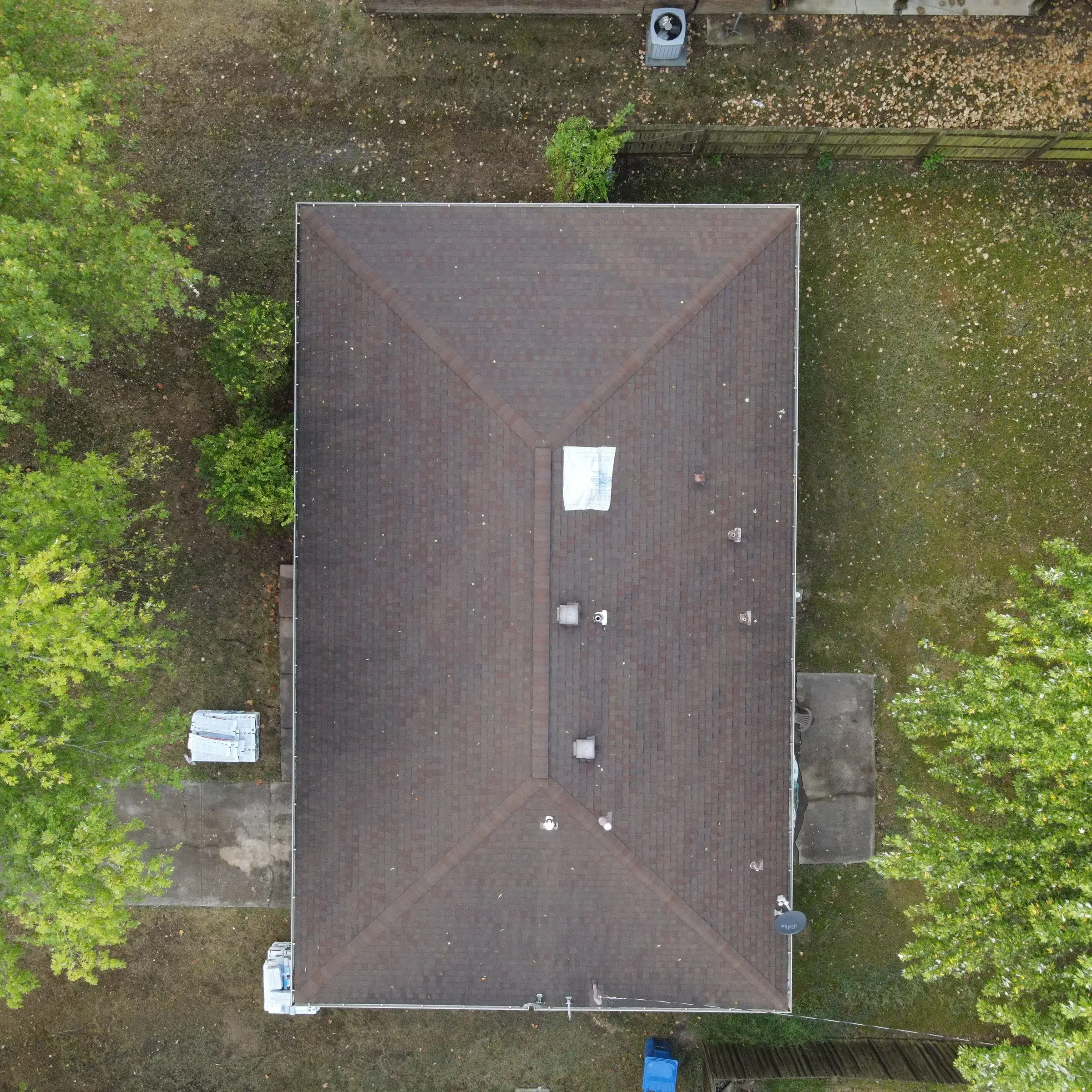 Overhead drone view of a long building with a brown hipped shingle roof, scattered leaves, and a white sheet lying near the center of the roof.
