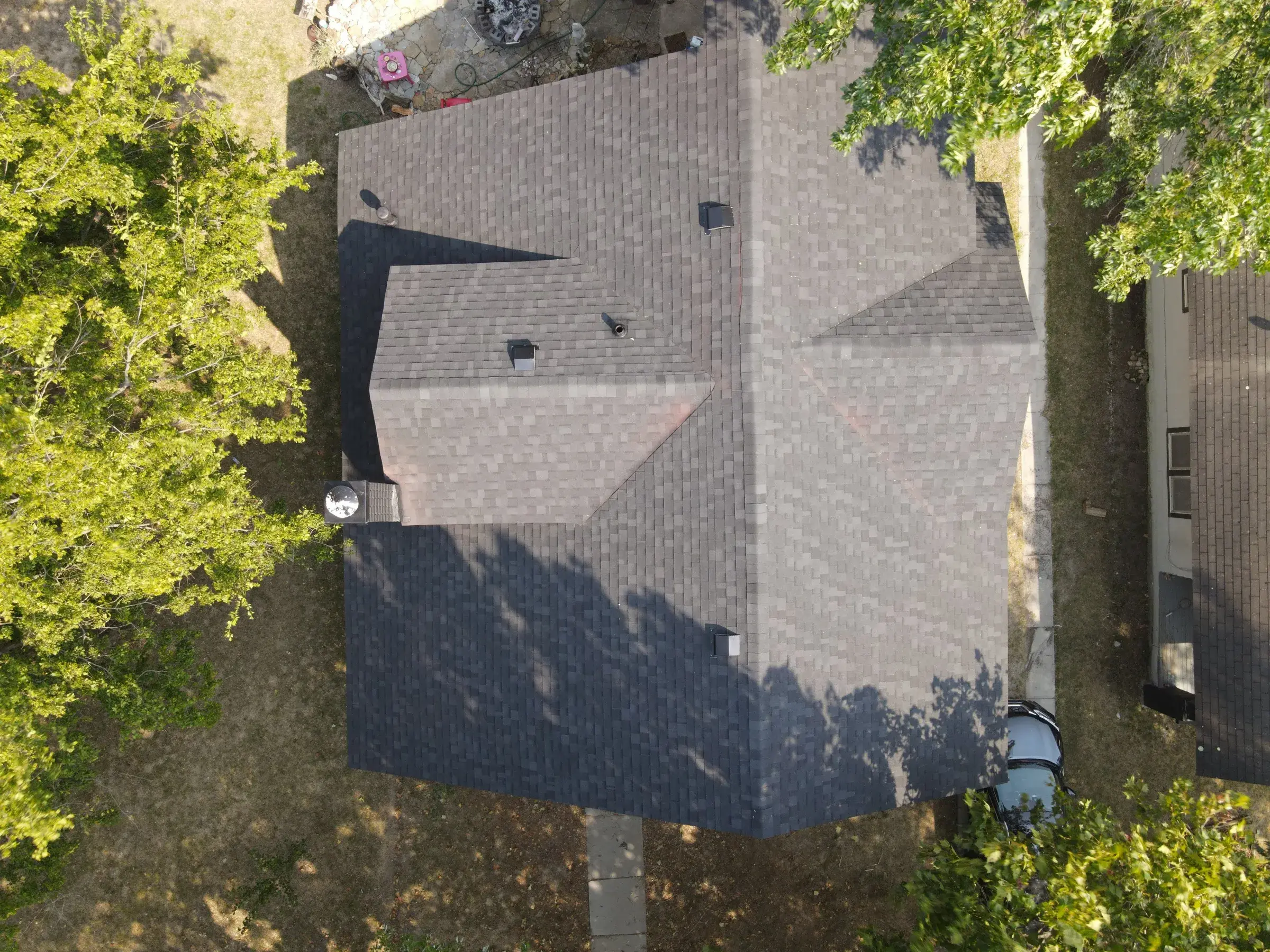 Overhead drone view of a house with a brown architectural shingle roof cut by hips and valleys, with tree shadows falling across the surface.