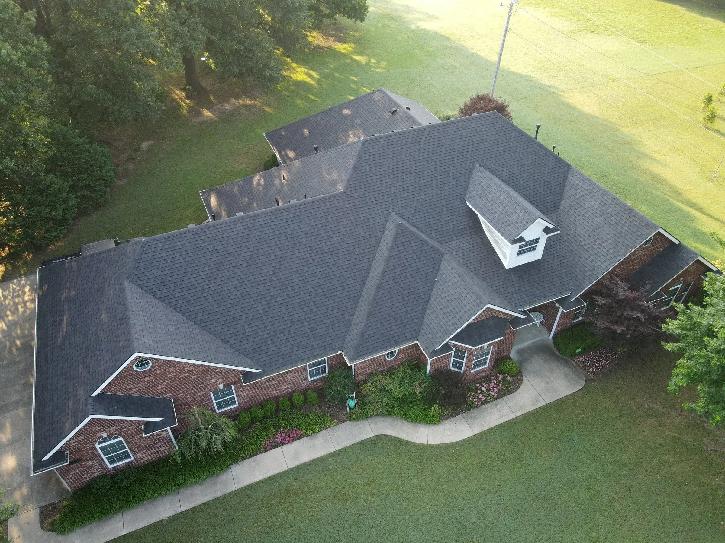 An aerial view of a large brick ranch home with a newly installed charcoal shingle roof, surrounded by mature trees and a green lawn in warm evening light.