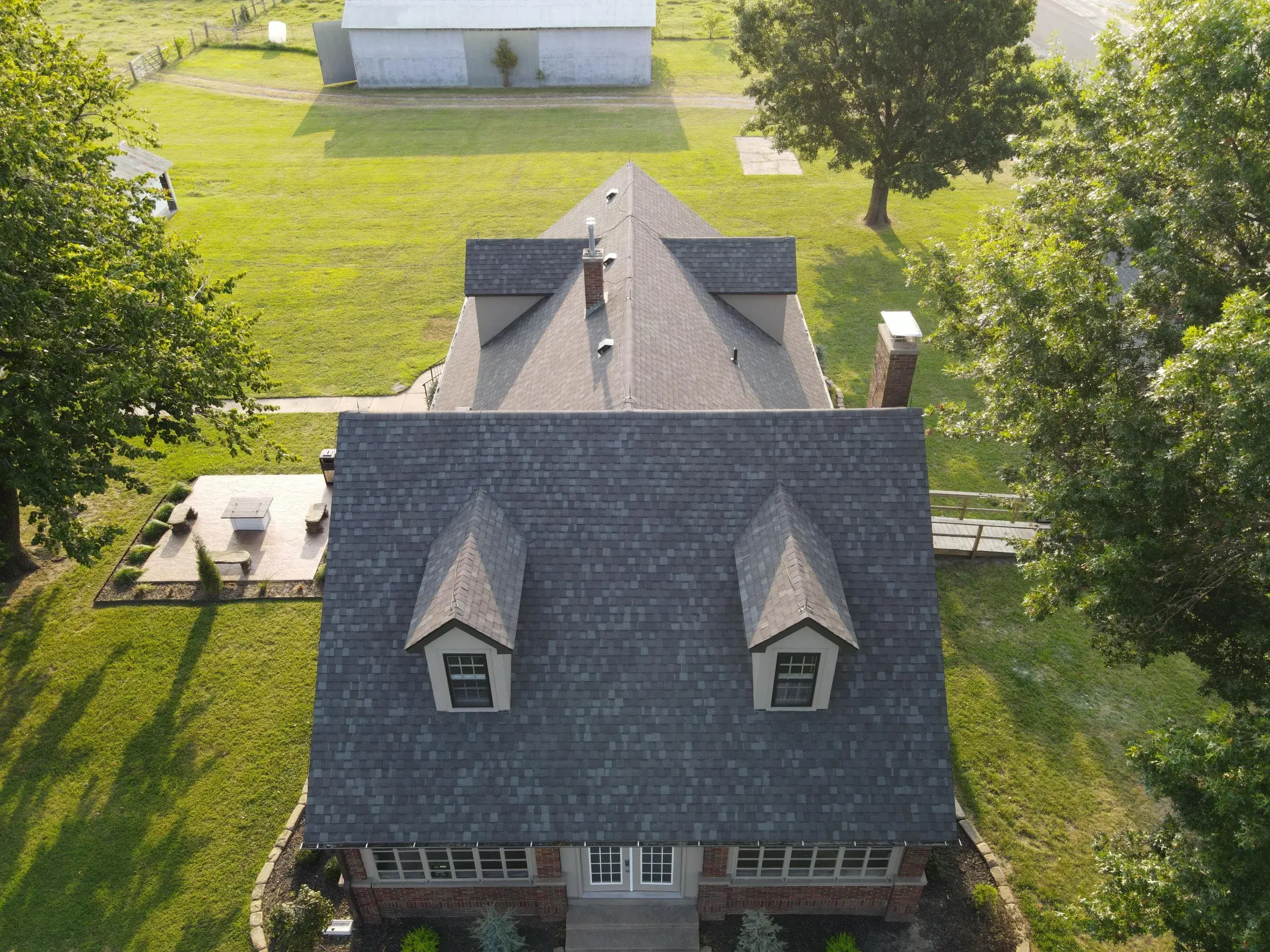 Aerial view of a brick home with a dark gray architectural shingle roof and two white dormers, lit by low golden sunlight and surrounded by mature trees and green lawn.