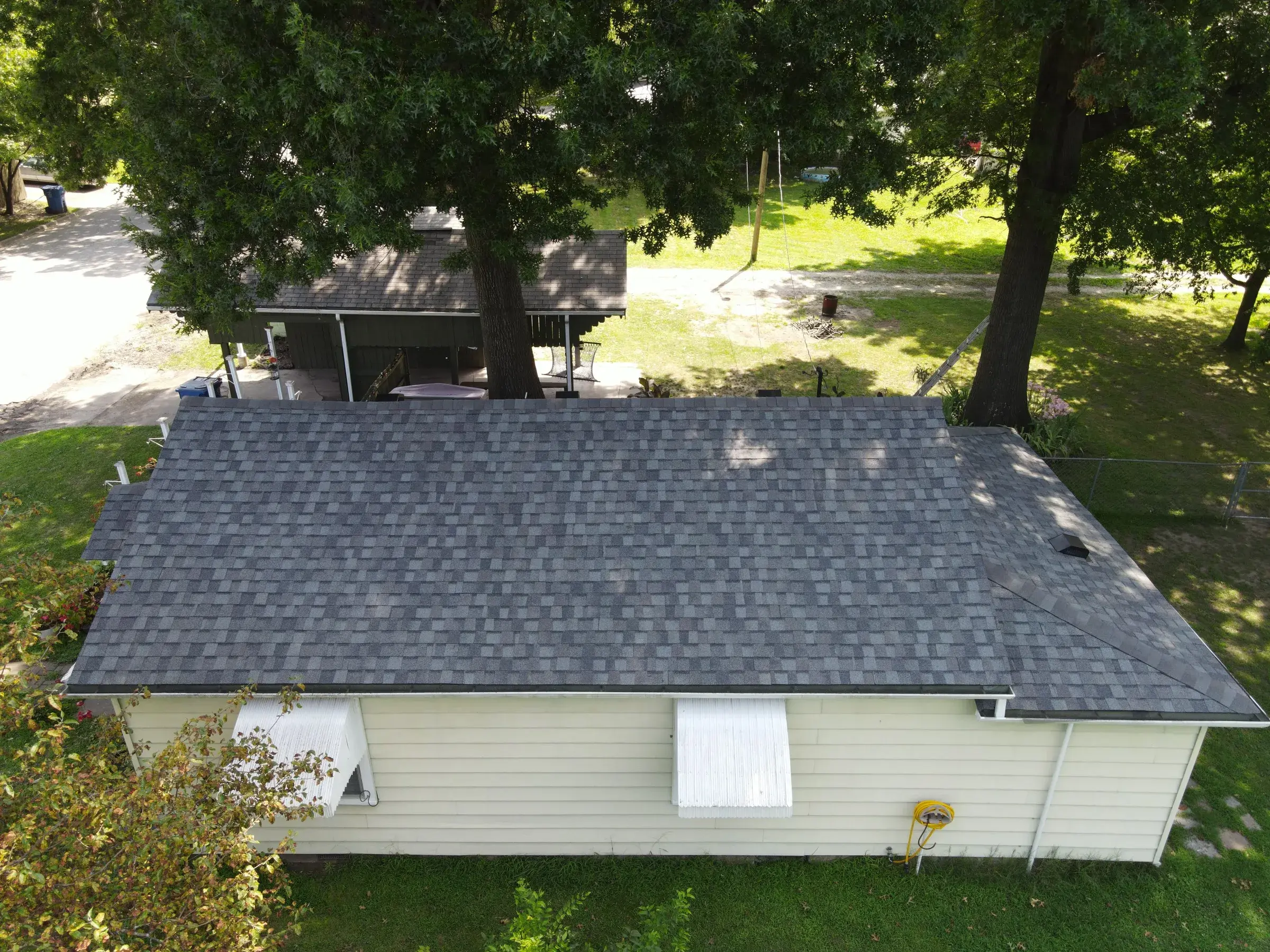 Aerial view of a small single-story house with a newly shingled gray gable roof, shaded by mature trees.