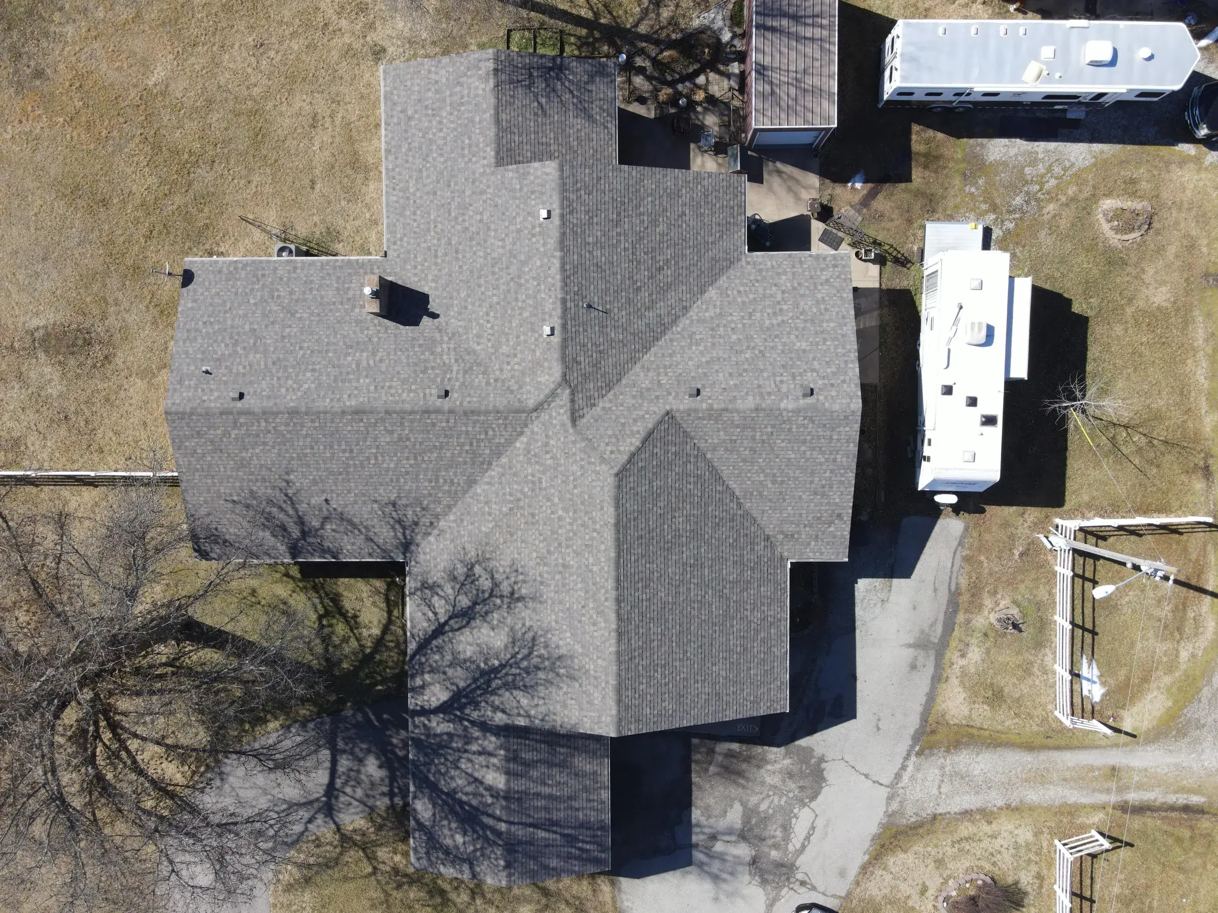 Overhead drone view of a large home with a clean light gray dimensional asphalt shingle roof, with two white travel trailers parked alongside on a gravel drive.