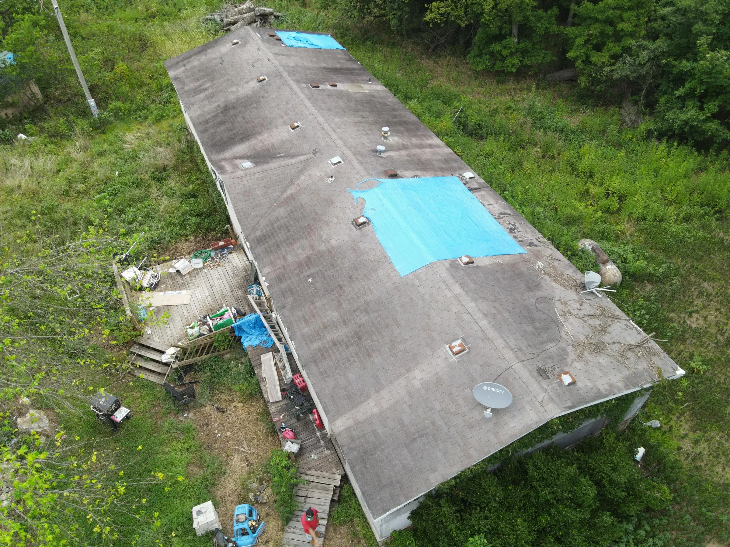 Aerial view of a long low-slope roof with two blue tarps covering damaged sections, surrounded by dense green brush.