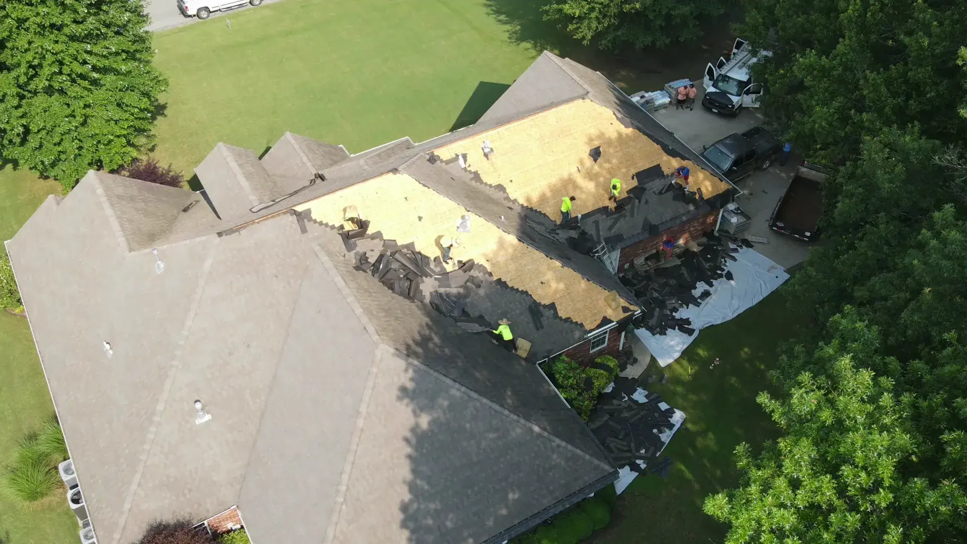 Aerial view of a large home with half its roof stripped to bare wood decking while a roofing crew works, with old shingles piled on tarps below.