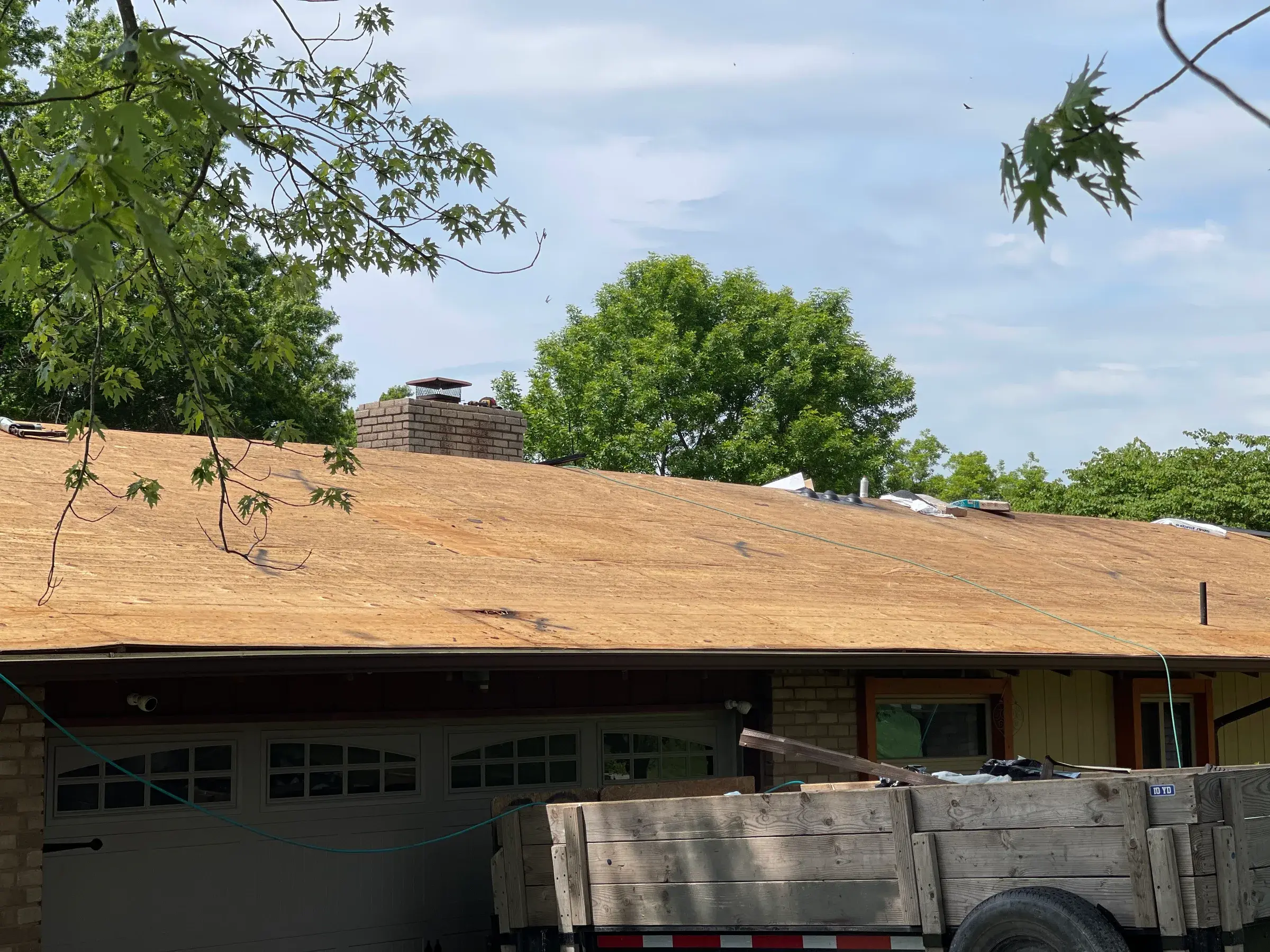 A brick home with its roof stripped down to bare wood decking, with a loaded dump trailer parked in front.
