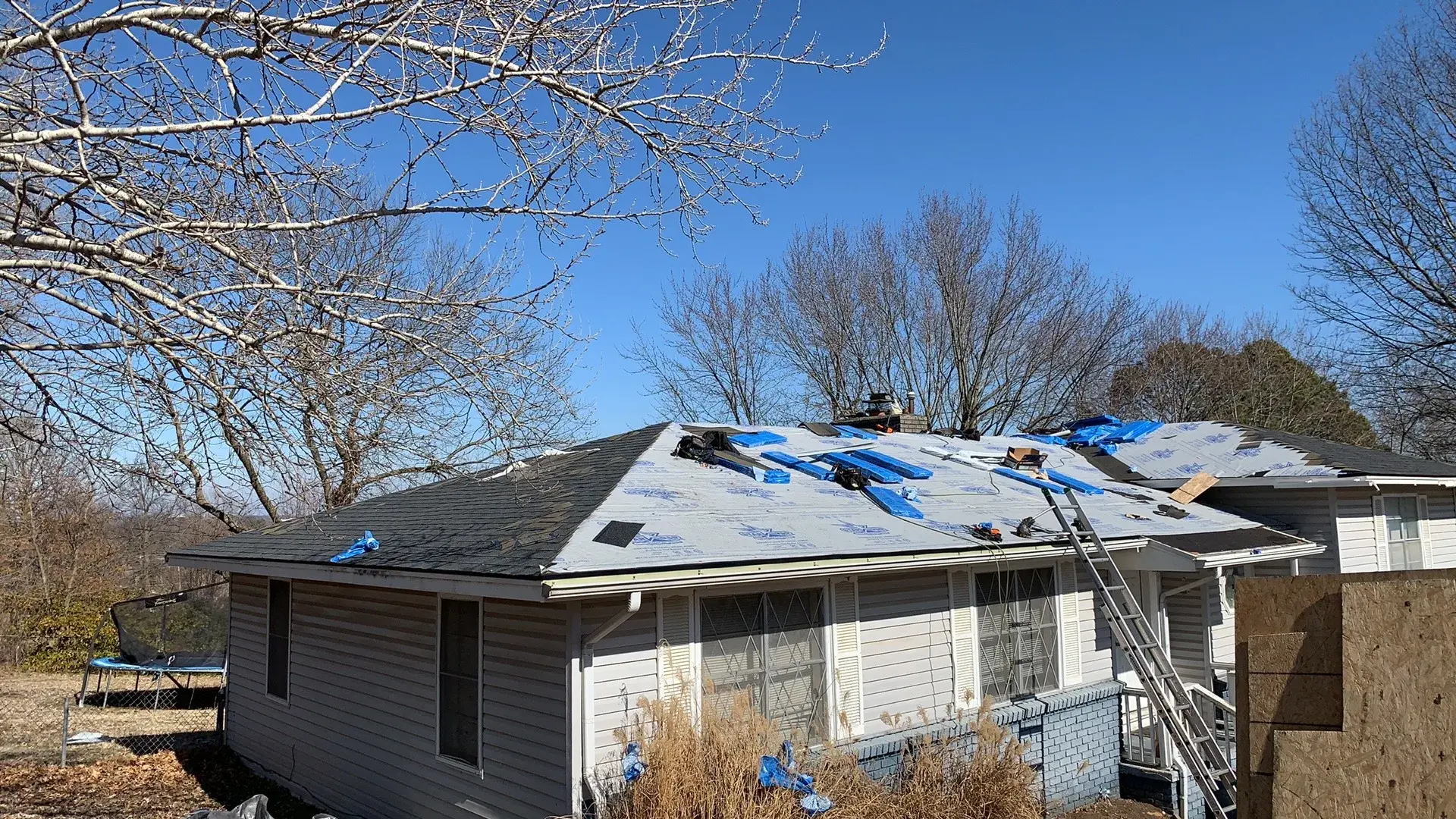 A single-story house mid-roof-replacement, with the old shingles stripped off, synthetic underlayment covering the deck, shingle bundles staged on the ridge and an extension ladder set against the eave.